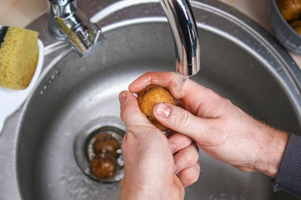 A man washes raw potatoes in sink under water to bake them in foil in the oven. — Fotografia de Stock