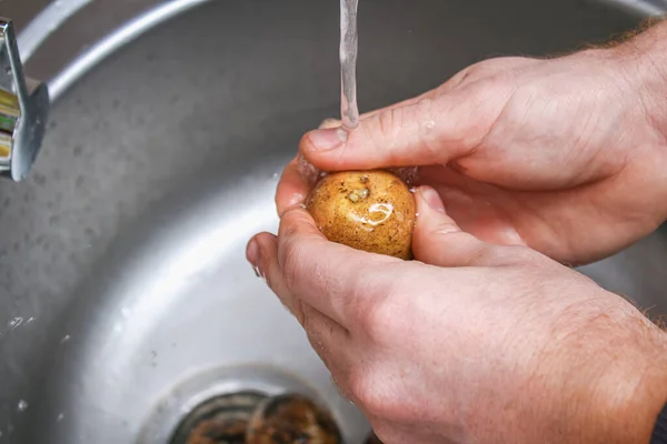 Человек моет сырой картофель в раковине под водой, чтобы испечь его в фольге в духовке. — стоковое фото