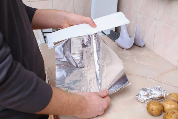 Um homem rasga o papel alumínio do dispensador de plástico para embrulhar batatas nele e assá-los no forno. — Fotografia de Stock