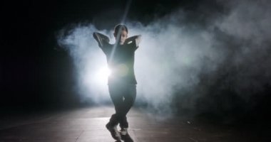 Genç bayan dansçı strop ışığı ve duman arka planıyla ağır çekimde hip hop dansı yapıyor.