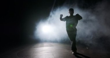 Genç bayan dansçı strop ışığı ve duman arka planıyla ağır çekimde hip hop dansı yapıyor.
