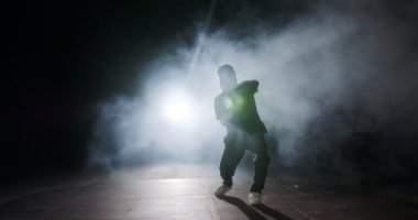 Genç bayan dansçı strop ışığı ve duman arka planıyla ağır çekimde hip hop dansı yapıyor.