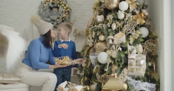 Matka a dítě zdobí vánoční stromek. Máma a pětileté dítě v modrých šatech zdobí vánoční stromek doma, dávají zlaté hračky na větve. — Stock video