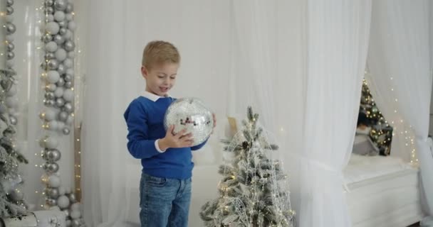 Веселый пятилетний блондин в синей одежде держит большой бал на елке на фоне яркой спальни, украшенной елкой. Новый год. Сочельник, сюрприз. — стоковое видео