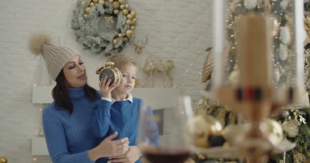 A mother with her son in blue clothes at a table, against the background of a Christmas tree, turn the clock in their hands, hug, listen to the ticking of the arrows. The clock is approaching twelve. — Stock Video