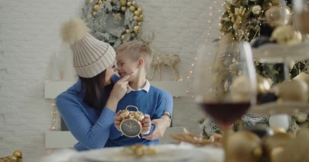 Одинокая мать и ее сын за праздничным столом, на фоне елки, поворачивают часы в руках, обнимаются, они счастливы. Часы приближаются к двенадцати. — стоковое видео