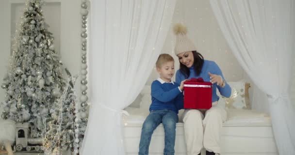 Familie vakantie. Vreugdevolle moeder en zoon begroeten Kerstochtend thuis op het bed naast een versierde kerstboom en houden een geschenk vast. Gelukkig gezin van 2 personen brengen Kerstmis samen thuis door. — Stockvideo