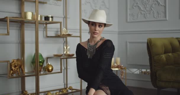 A luxurious rich lady of 30 years old in an expensive interior, dressed in a black dress and a white hat, poses in front of a mirror. — Vídeo de Stock