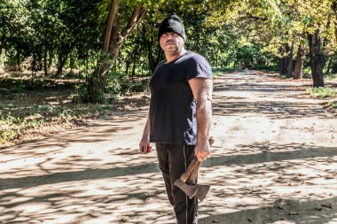 Şapkalı ve elinde balta olan bir adam ormanın ortasında yolun ortasında duruyor..