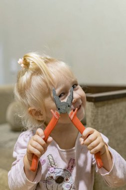 Küçük bir kız yüzünün önünde pense tutuyor ve bir göz kırpıyor gülümsüyor.