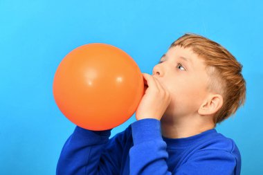 Çocuk mavi arka planda turuncu bir balon şişiriyor..