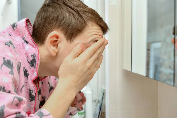 A young man washes his face with water from the tap in the washbasin and wipes his face with his hands in the bathroom.