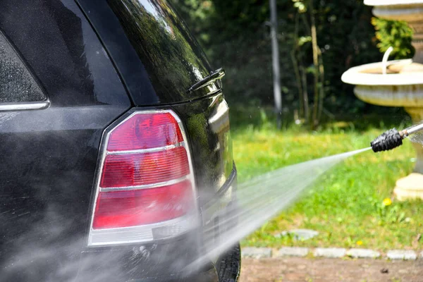 A driver washes a black car with a high-pressure water jet at his home.