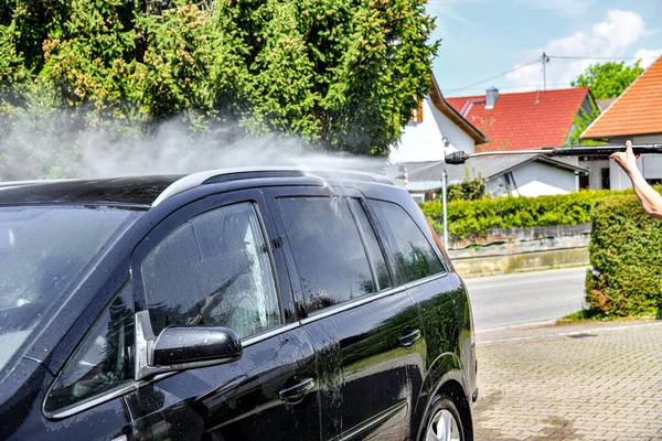 A driver washes a black car with a high-pressure water jet at his home.