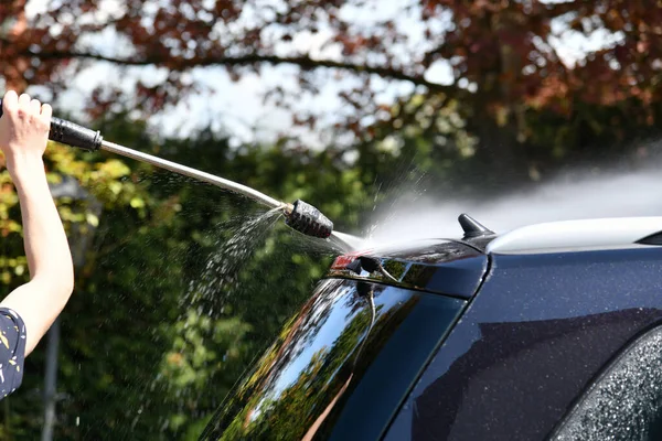 A driver washes a black car with a high-pressure water jet at his home.