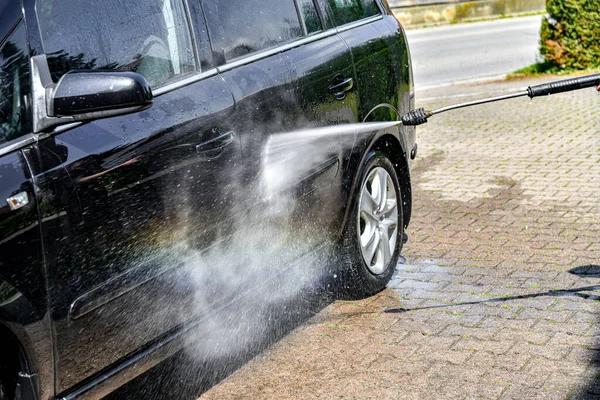 A driver washes a black car with a high-pressure water jet at his home.