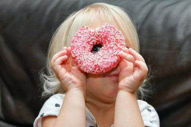 Küçük neşeli bir kız yüzünün yanında kırmızı bir çörek tutuyor ve ona bakıyor..