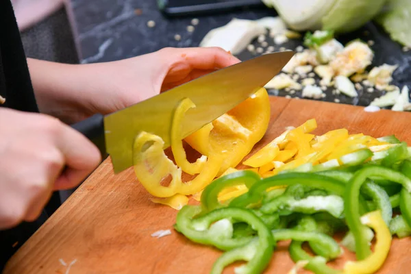 A woman in the kitchen cuts yellow and green peppers into small pieces.