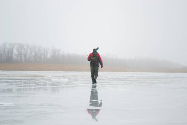Novopskov, Ukrayna - 2 Ocak 2021: Bir balıkçı kışın elinde matkapla havuzda. Sisli bir sabahta kırsalda kış balıkçılığı. Ahenk ve barış kavramı. Balıkçı kameraya arkasını döndü ve uzaklaştı.
