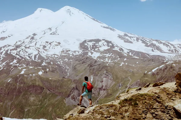 Человек с красным рюкзаком стоит на скалистой горе и смотрит на снежную вершину Эльбруса. Стоковое Фото