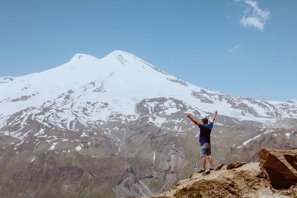 Man staan op rotsachtige berg en kijk naar besneeuwde piek van Elbrus. Stockafbeelding