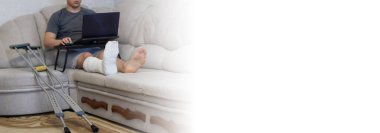 Adam kanepede yatıyor ve bacağında bandajla dizüstü bilgisayarlarla çalışıyor..