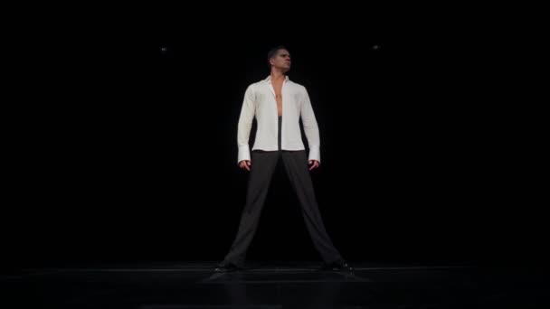 一个严肃的年轻人，穿着没有扣子的白衬衫站在黑色舞台上，两腿分开，舞姿迥异，头从左向右，女人穿着短裙跟在后面跳华尔兹。. — 图库视频影像