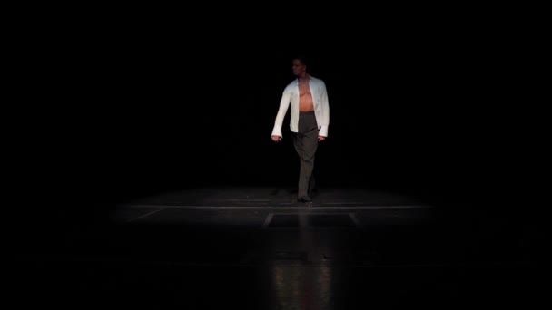 Ballroom male dancer on stage in white shirt,naked tanned torso,moving from darkness to floodlight. making swing with his hand,slow steps in circle.Solo performance fragment.Distance shot,front view. — Stock Video
