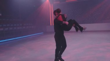Artistik patinajcılar buz pateni pistinde kaldırış yaparken dizlerinin üzerine çöküp yavaşça dönen kadını tutan adamı neon kırmızısı ışıkta buza yatırıyor. Buz tiyatrosu konsepti, ağır çekim.