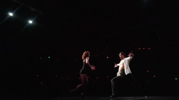 Mężczyzna tancerz podnosi kobietę z góry nogami do góry nogami, podnosi ją do tańca na ramieniu od tyłu, obraca się z nią w pięknej pozie, ramiona wyciągnięte.Aparat latać przez taniec para na ciemnej scenie. — Wideo stockowe