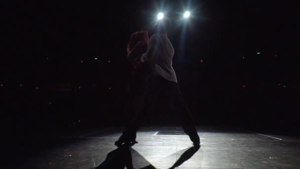 El hombre abraza apasionadamente a la mujer inclinada hacia atrás, los pasos de tango girando, entran en la proa. Pareja de baile de salón en el escenario iluminado, vista trasera desde el escenario contra el fondo oscuro del auditorio. Punto de baile final. — Vídeo de stock