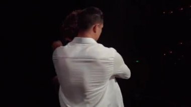 Balo dansçıları sahne performans parçacığı taşkınlarını eşleştiriyor. Erkek partner, kadının belini destekliyor, dizleri göğsüne kadar kıvrılıyor, onu indiriyor, kamera geri çekiliyor, kamera döndükten sonra hareket ediyor.