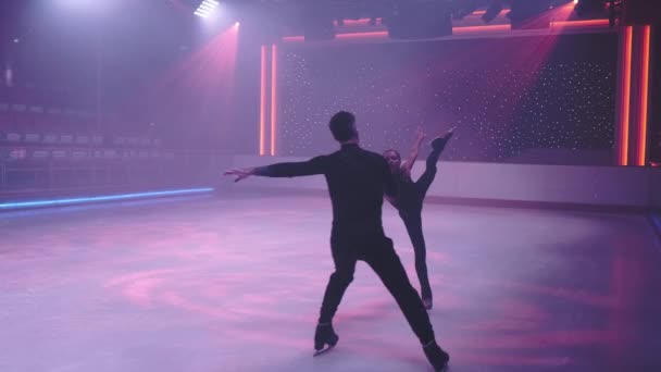 Figure skaters pair on ice rink in neon light skating by camera,man moves backwards holding woman hand,performing arabesque,leg and arm raised parallel high up behind.Slow motion. Camera tracking — Stock Video