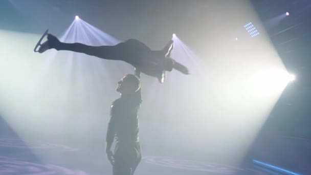 La pareja de patinadores gráficos realiza un levantamiento de techo rotatorio estacionario en reflectores. El hombre sostiene a la mujer paralela al hielo en su mano recta, sus piernas en división longitudinal. Ensayo.Cerrar siluetas. — Vídeo de stock