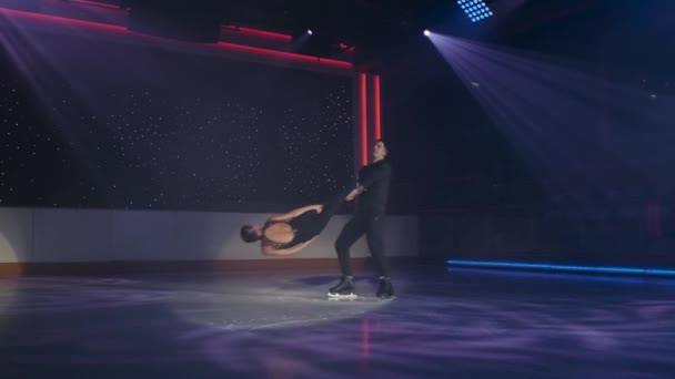 Par de patinadores artísticos realiza elevação rotativa estacionária em holofotes. O homem segura a mulher por ambos os tornozelos com os braços cruzados, girando, ela paira sobre o gelo com os braços endireitados ao longo do corpo. Ensaio — Vídeo de Stock