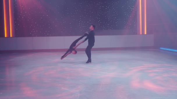 Figure skaters pair on ice performs stationary rotational support,prohibited element. Woman in split upside down,hips angle 180, man in crouch holding her with straight hands by arm,leg.Slow motion. — ストック動画