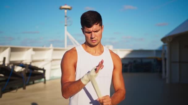 Athletic attractive man puts on hands wraps to do box outside onboard cruise ship — Stockvideo