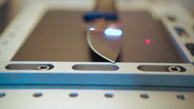 Hunting black knife on laser machine work platform in engraving process,laser beam moving, camera accelerated zoom in, drawing feathers pattern on metal blade surface, close-up front view. Workshop