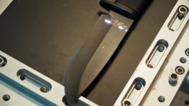 Process of laser engraving of hunting knife surface with wide blade, drawing pattern of feathers, close-up, top view. Working with laser on metal concept in workshop.Ray movement from handle to middle