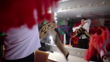 Pasadoble dancer in white shirt in dressing room in front of mirror looks at his reflection,slowly rotating throws bullfighters vest over shoulders.Costumes,wigs and feathers,head mannequins around