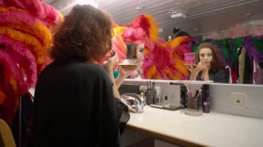 Beautiful woman actress applies eye makeup,holds compact eye shadow palette with mirror in front of her,sits in dressing room at table. Rear side view,camera zooming.Bright dance costumes,feathers