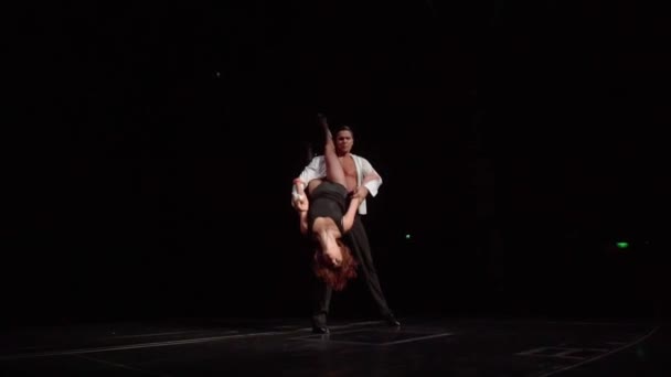 拉丁美洲的交际舞舞伴在黑暗的舞台上 男人倒挂着美丽的长腿女人 弯腰站在后面 转过身来 摆出性感的姿势 遥视相机飞驰而过 动作缓慢 — 图库视频影像