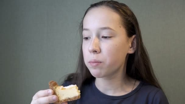 Uma adolescente com aparelho come um pedaço de bolo de queijo cottage, de pé em casa junto à janela contra uma parede verde, e olha para o lado. Close-up. Conceito. 4K. — Vídeo de Stock