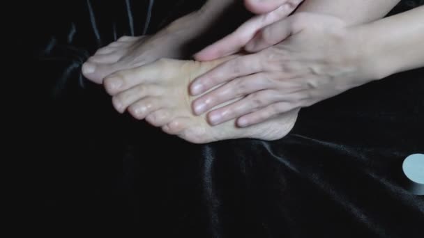 Una mujer masajea una pierna y dedos de los pies cansados y desnudos, sentados en la cama sobre una sábana negra. Concepto. Primer plano. Sin rostro. Aislado. 4K. — Vídeo de stock