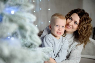 Çekici bir çocuk ve genç annesi evdeki güzel bir Noel ağacının yanında oturmuş kameraya bakıyor ve gülümsüyorlar. Yakın plan. Mutlu yıllar konsepti. Portre. Yatay görünüm