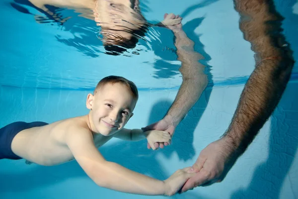 Retrato de un niño feliz bajo el agua, con las manos de sus padres bajadas al agua. Él toma de la mano y sonríe. Clases de natación. Estilo de vida saludable. Primer plano. Orientación horizontal de la foto. — Foto de Stock