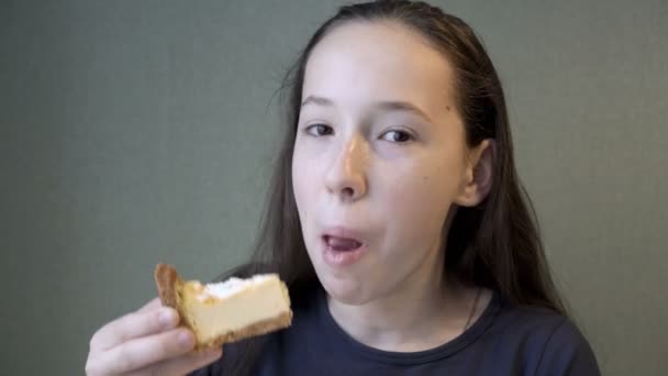 Uma adolescente bonita com aparelho de metal nos dentes sorri, olha para a câmera e começa a comer um pedaço de bolo de queijo cottage, em casa junto à janela contra uma parede verde. Close-up. 4K — Vídeo de Stock