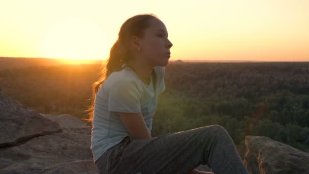 Ein nettes, nachdenkliches Teenager-Mädchen sitzt und träumt auf einem Berg vor der Kulisse eines Sonnenuntergangs und einer wunderschönen Landschaft. Schöne Aussicht auf das Tal bei Sonnenuntergang an einem Sommertag. 4K — Stockvideo