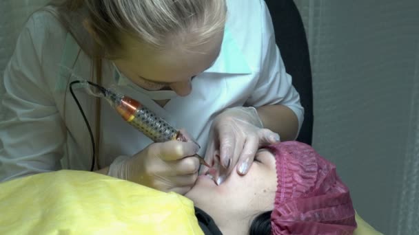 Жінка-косметолог у білому пальто робить татуювання на губах для дівчини-клієнта в салоні краси. Перманентна процедура макіяжу губ. Крупним планом. 4-кілометровий — стокове відео