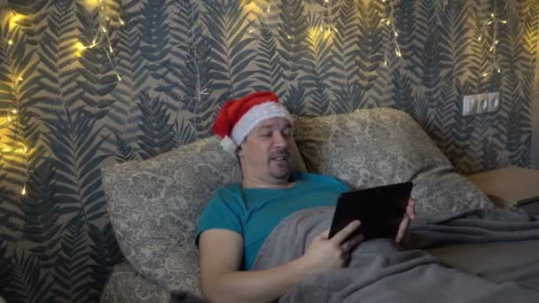 Un hombre adulto enfermo se comunica a través de un enlace de vídeo a través de una tableta mientras está acostado en casa en una cama con un sombrero de Santa Claus. El concepto de celebrar la Navidad durante la pandemia del coronavirus. 4K — Vídeos de Stock